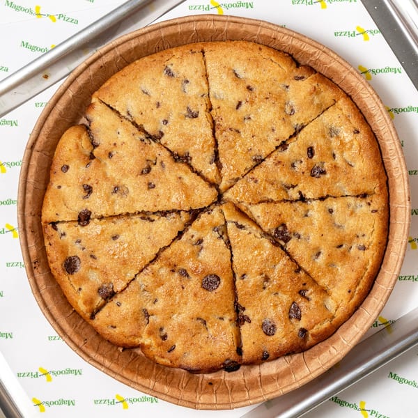 A large, round chocolate chip cookie pizza, sliced into eight wedges, in a brown paper pan.