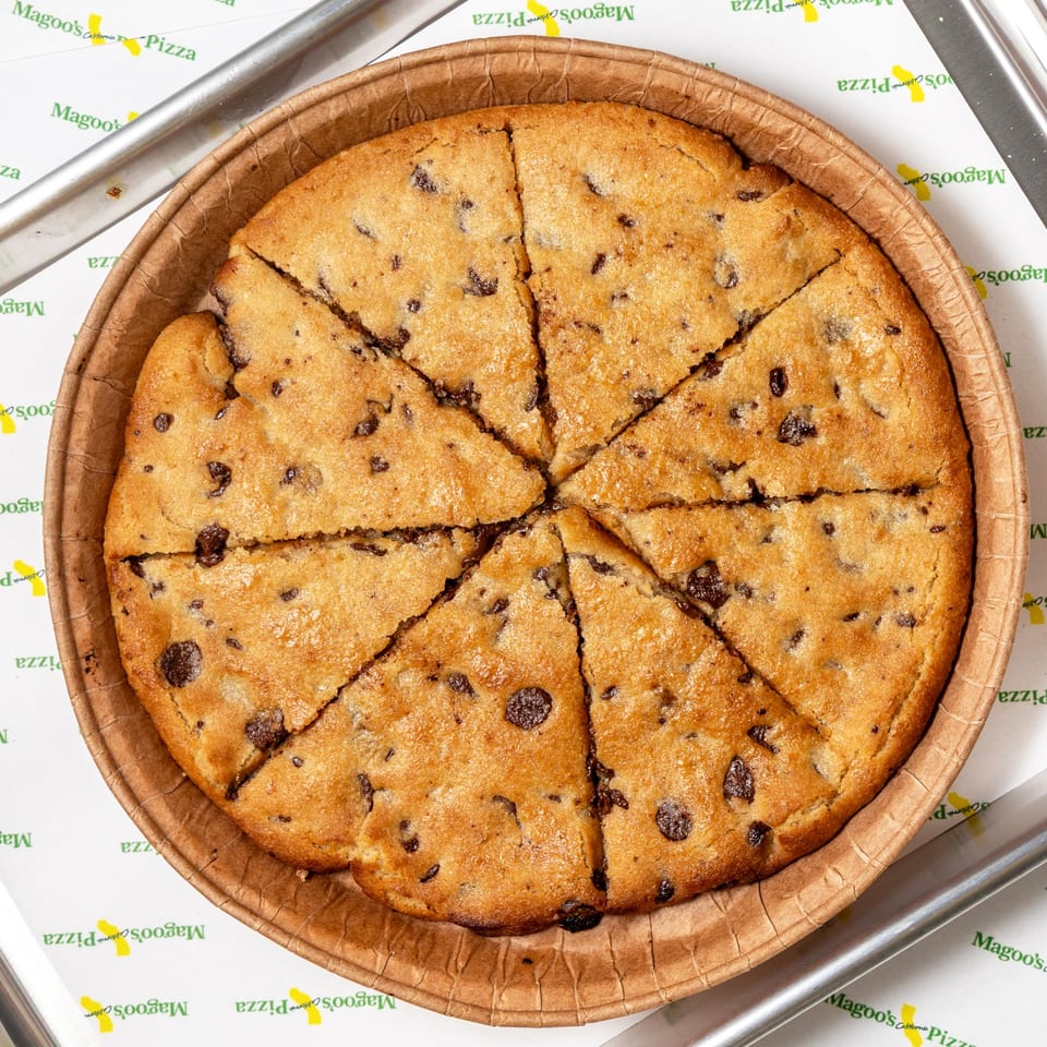 A large, round chocolate chip cookie pizza, sliced into eight wedges, in a brown paper pan.