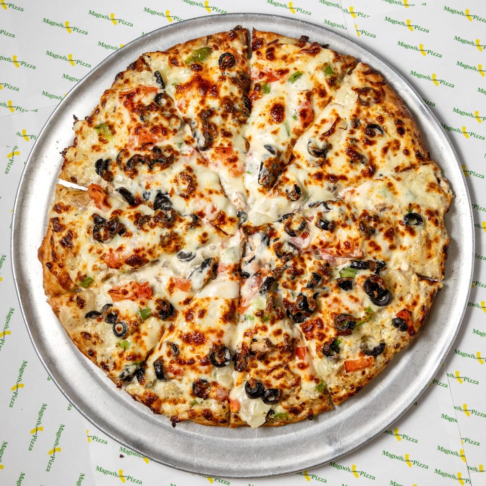 A whole Magoo's California Pizza, sliced, with melted cheese, olives, and vegetables on a metal pan.