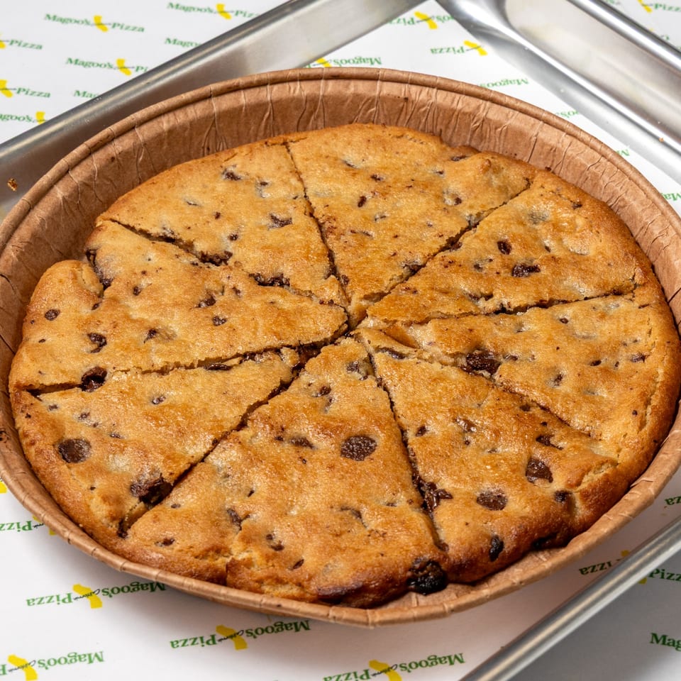 A large chocolate chip cookie, cut into eight slices, in a round brown paper container.