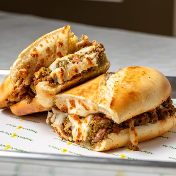 Two halves of a cheesy steak and pepper sub sandwich on Magoo's Pizza branded paper.
